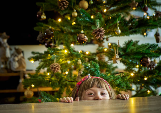 A curious child peeking over a festive table, with a Christmas tree in the background, captures the magic, warmth, and joy of the holiday season. A girl swung with anticipation lights up the scene