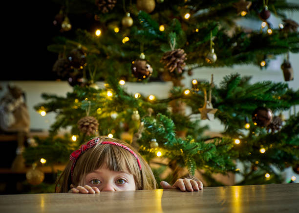istock A innocent child with a red headband is peeking over a table in front of a decorated Christmas tree. The adorned tree creates a festive atmosphere. The girl conveys festivity and anticipation concept 2189756332