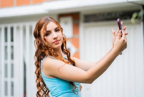 young woman with long wavy hair outdoors taking a selfie on her cell phone and looking at the camera