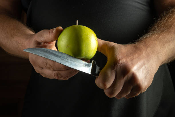 istock Hands grip a sharp knife poised over a vibrant green apple, emphasizing the tension of preparation. The soft glow of the kitchen casts a warm ambiance, highlighting the act of cutting with care 2189772753