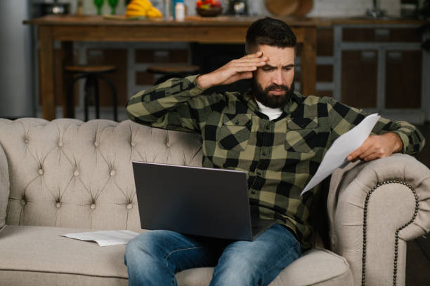 istock Young man with beard analyzing budget or utility bills, looking worried sitting at home on the sofa 2189776808