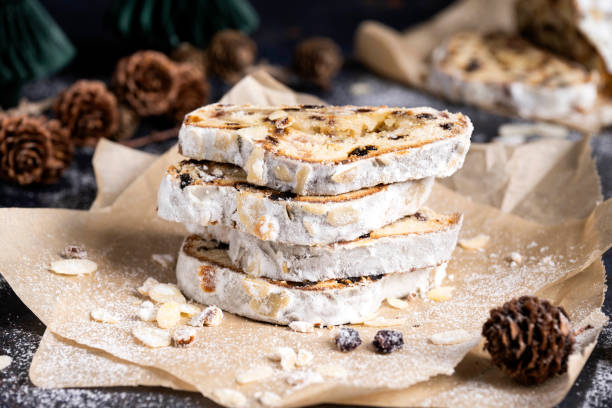 Sliced German Christmas fruit cake known as Stollen cake on parchment paper. There are Christmas decorations in the background.