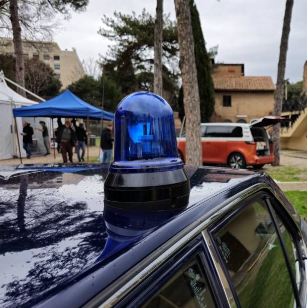 A police car with a blue emergency light in focus, set in a rural area with tourists, tents, and vehicles in the background, suggesting security and vigilance. Rome, Italy - December 13, 2024