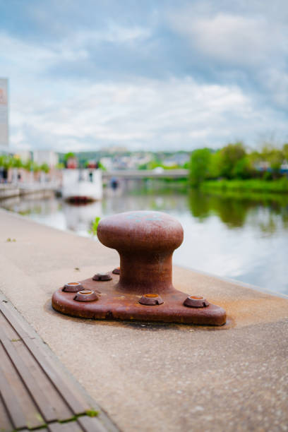 Rusty metal bollard securely fixed on a waterfront, with a blurred view of a river, urban structures, and greenery set against a cloudy blue sky.