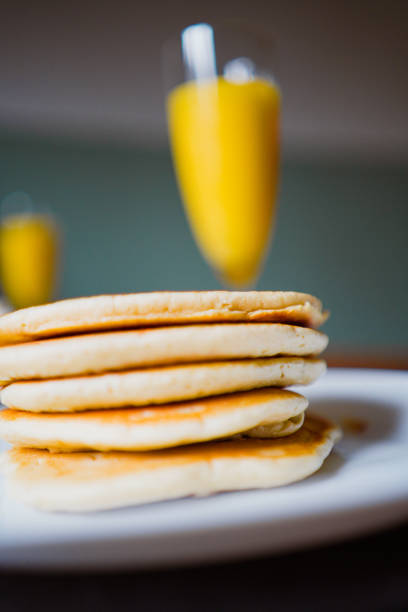 Close-up view of a stack of golden brown, freshly cooked pancakes on a white plate, paired with orange juice in elegant stemmed glasses blurred in the background, conveying a cozy breakfast atmosphere