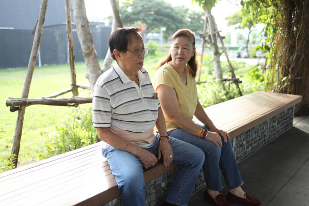 The senior Asian man, wearing a lumbar support belt, is sitting with his wife in the park during the daytime.