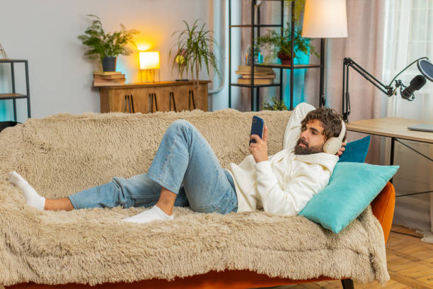 istock Happy overjoyed young man in wireless headphones using smartphone relaxing sitting on sofa at home 2189836496
