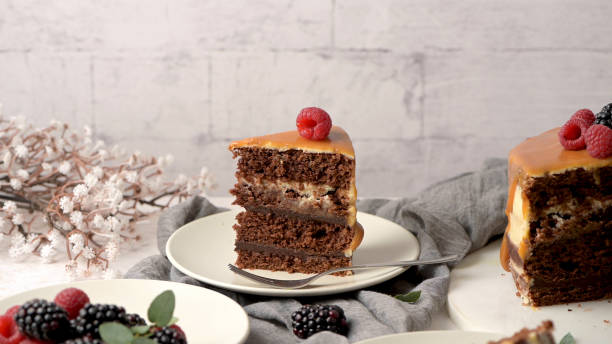 istock Chocolate cake slice with caramel frosting and a raspberry on top, surrounded by fresh berries and a floral arrangement, creating a cozy dessert scene 2189841342