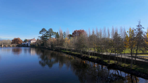 istock Panoramic view of a river with a stone bridge, a historic building, and a tree-lined path along the shore under a clear blue sky 2189843175