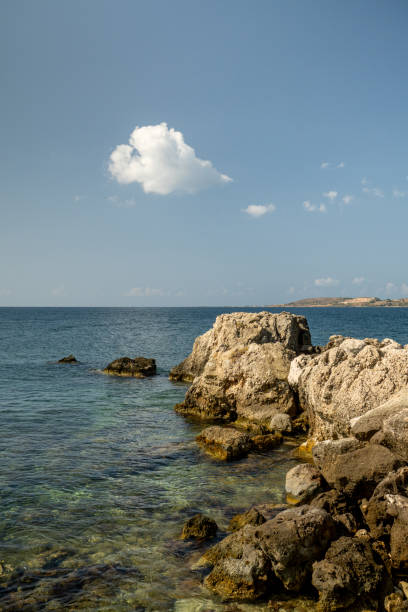 A rocky seascape, adorned with jagged cliffs and boulders, stretches out under the vast, open sky. Above, a solitary cloud drifts lazily, casting shifting shadows over the rugged terrain below. The interplay of light and shadow, rock and water, creates a serene yet dramatic tableau, a testament to nature's raw beauty.