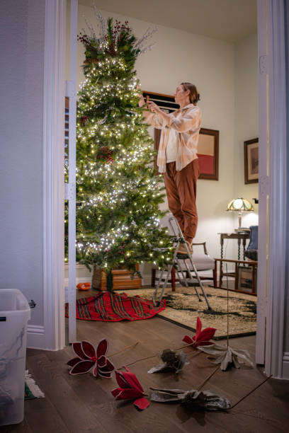 woman in pajamas stands on step stool while decorating Christmas tree