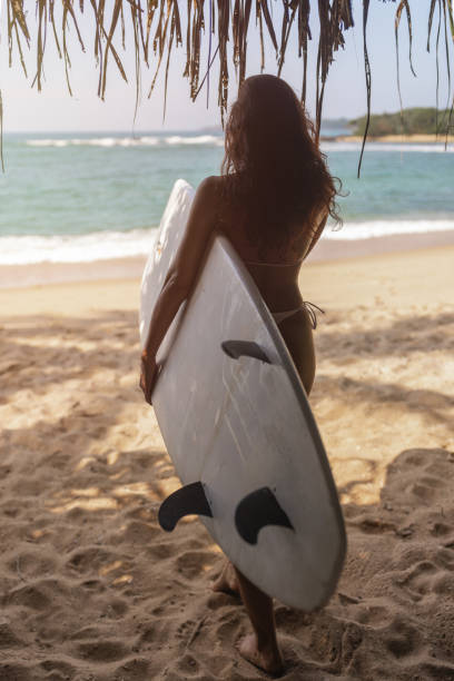 istock Woman with Surfboard Standing Under Palm Shade on a Tropical Beach 2189872453