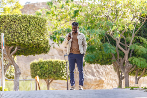 istock Young man looking at his phone while walking through a beautifully landscaped park in daylight 2189900991