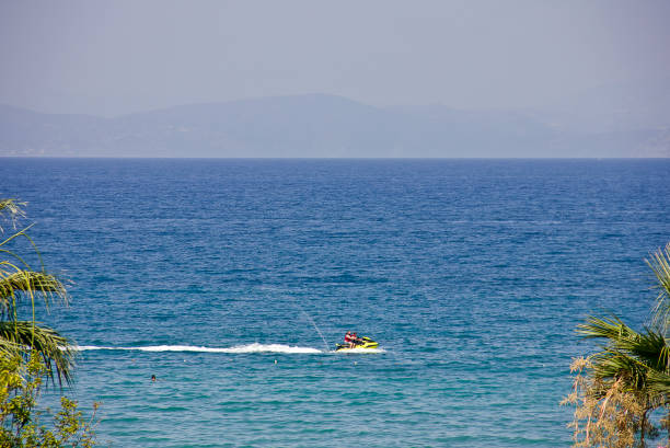 Aydın, Turkey - July 19, 2024: People enjoying near the sea at beach on summer holiday. Some of them swimming and others are parasailing on Aegean sea, you see town of Kuşadası in Turkey on a sunny summer day, View Of Kusadasi Skyline At Aegean Coast, Turkey.