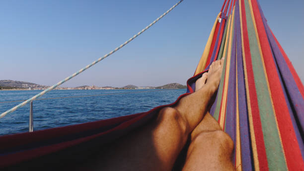 Man Relaxes in a Hammock on a Sailboat,his Legs Crossed at the Ankle as he Enjoys the Bright Sunny Day. The Calm Sea and Clear Sky Enhance the Peaceful Atmosphere,Creating a Perfect Moment of Tranquility on the Water