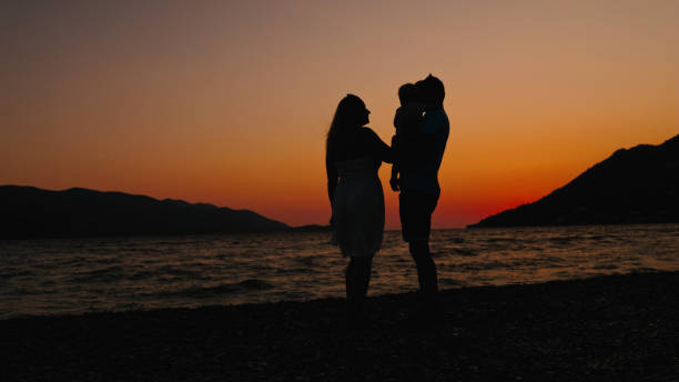 Silhouetted Against the Warm Hues of a Setting Sun,a Mother and Father Stand at the Coastline,Holding their Little Child in their Arms. The Serene Moment Captures the Essence of Family Love and Connection Amidst the Tranquil Beauty of the Beach at Sunset