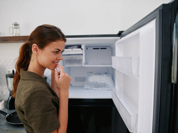 istock Woman smiling with teeth looking into camera in kitchen at home opened freezer empty with ice inside, home refrigerator, defrosted, view from back, stylish interior. 2189944940