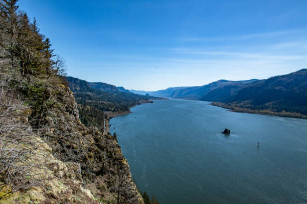 Far in the distance, a young solo female hiker stands atop a rock overlooking a mighty river. A mountain range rises up on the other side. The view is magnificent.