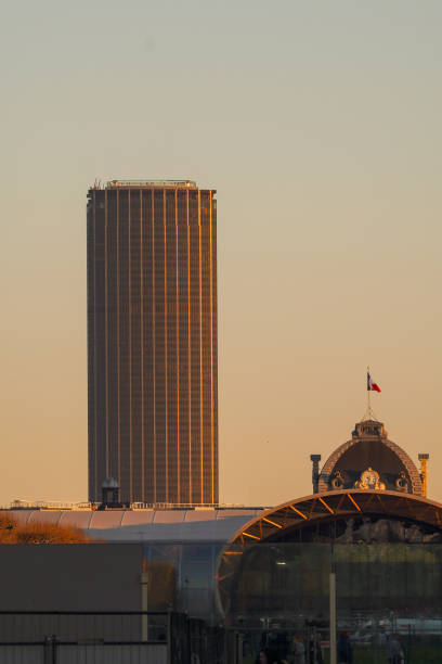 The Montparnasse Tower rises above Parisian rooftops during golden hour. A blend of modern and traditional architecture under the warm hues of the setting sun.