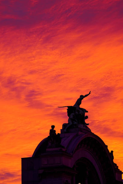 Dramatic silhouette of a statue with figures reaching skyward, set against a vivid red and orange sunset sky. A powerful and artistic architectural scene.K
