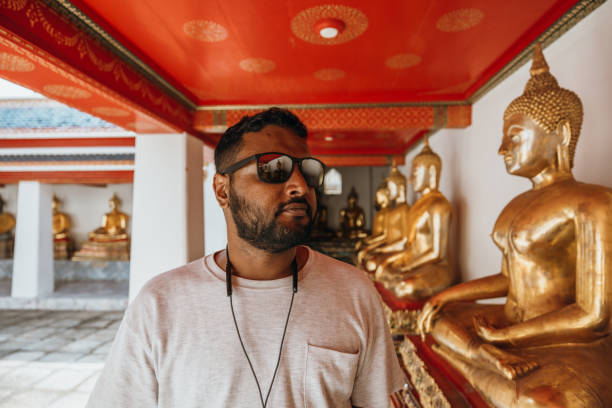 Man in white shirt walks past golden Buddha statues in a temple. Reflective visitor in Asian shrine. Exploring cultural heritage and religious art.