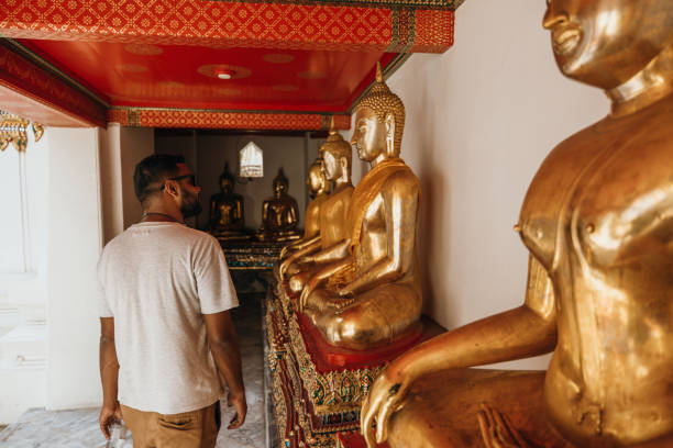 Man in white shirt walks past golden Buddha statues in a temple. Reflective visitor in Asian shrine. Exploring cultural heritage and religious art.