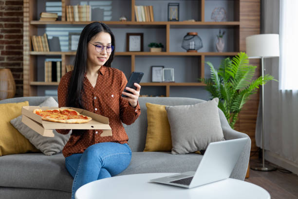 istock Asian woman enjoying pizza at home while browsing on phone, sitting on cozy sofa. Modern lifestyle with technology. Relaxed casual atmosphere featuring a laptop and casual wear. 2190032227
