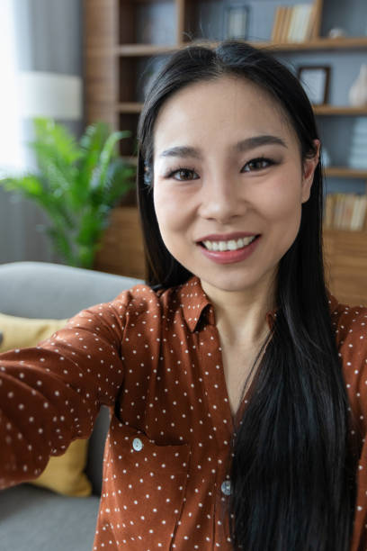 istock Asian woman taking selfie at home with smartphone, sitting on sofa, smiling while capturing happy moment during leisure time indoors, surrounded by cozy interior, showing personal lifestyle. 2190035671