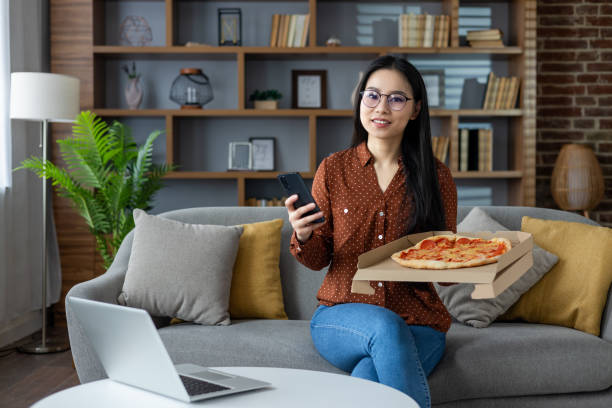 istock Asian woman enjoying pizza at home while using smartphone for casual relaxation time on weekend. Cozy living room with couch, laptop, and modern decor for a peaceful ambiance. 2190037246