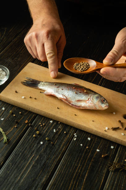istock Hands skillfully prepare a fresh fish on a rustic wooden board, sprinkling it with seeds and salt. The surrounding spices and herbs create an inviting atmosphere for culinary creativity. 2190037268
