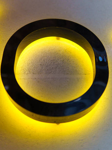 A circular LED light fixture emits a warm yellow glow against a textured wall. The design stands out with its sleek black frame, creating a modern aesthetic.