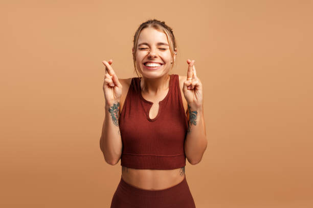 Smiling sporty young woman closing eyes, showing hope gesture with crossed fingers in studio Happy fitness young woman praying with crossed fingers on beige background, closing eyes. Emotion, sport, wellness concept dream believe tattoo stock pictures, royalty-free photos & images