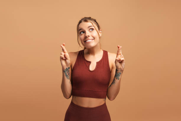Smiling fit woman crosses fingers, believing in good fortune, posing in studio Happy young sporty woman showing gesture with crossed fingers, posing, isolated on beige in studio. Emotion, sport, wellness concept dream believe tattoo stock pictures, royalty-free photos & images