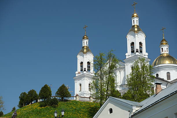 A church stands on a green hill, showcasing its stunning golden domes against a vibrant blue sky. The surrounding area features lush trees and blooming flowers, creating a serene atmosphere.