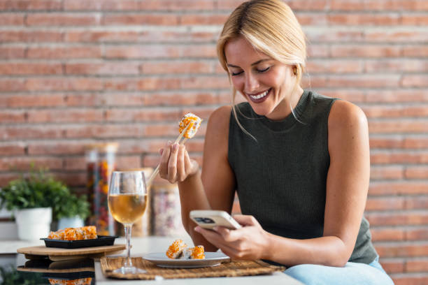 Pretty young woman eating sushi while using smartphone in the kitchen at home