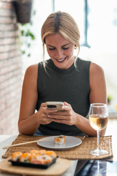 Pretty young woman eating sushi while using smartphone in the kitchen at home