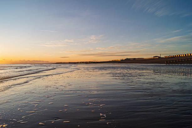A photograph taken of sunset at low tide on the beach at West Marina, St Leonards on Sea, East Sussex.  The image shows waves rolling gently in on the exposed sand. The colours of the sunset and setting sun are reflected in the sea and wet sand. The beach huts glow with the reflection of the sun.