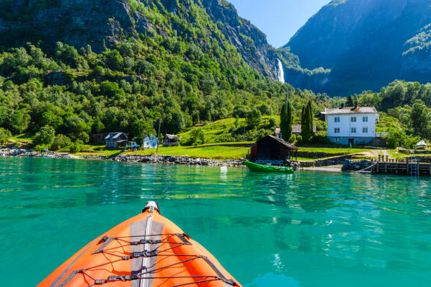 Paddling through serene turquoise waters, a kayaker enjoys the breathtaking views of rugged cliffs and lush greenery, with a picturesque village nearby and waterfalls cascading down.