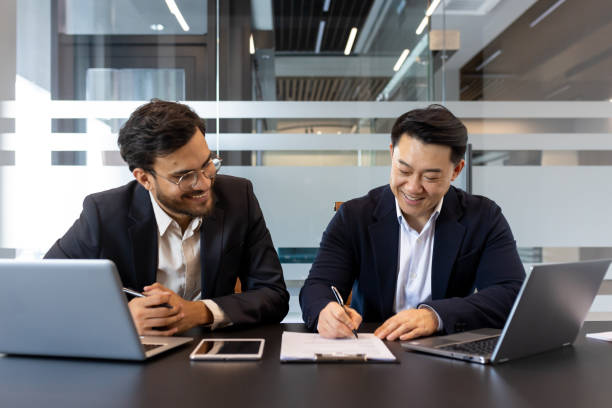 istock Two businessmen in a modern office environment signing a consent agreement while using laptops and a smartphone, showcasing teamwork and professional collaboration 2190094837