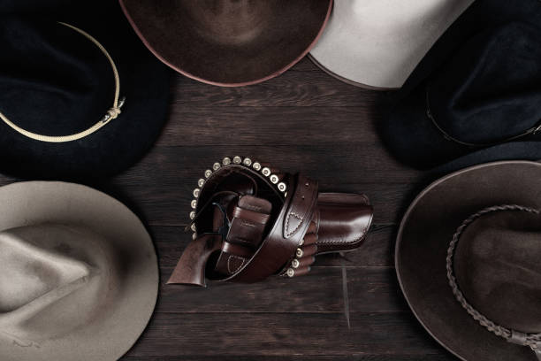 Gun with holster and gun belt with old west hats on wooden table. Top view.