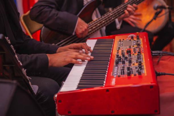Musicians Performing Together in a Live Orchestra Setting. Close-up of a musician playing a red color electronic keyboard during a live orchestra performance, accompanied by a bass guitarist in the background. The image captures the hands-on instruments, reflecting harmony, teamwork, and the essence of live music events