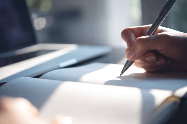 istock Close up of woman hand with a pen writing on notebook with laptop computer on office table. Business woman planning, making a note reminder. Student studying online, e-learning 2190147554