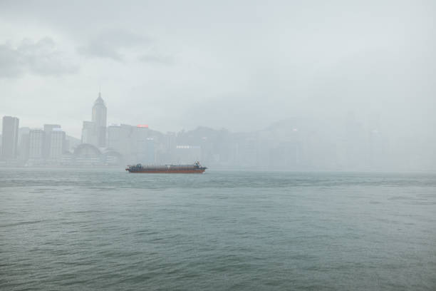 A cargo ship sails across Victoria Harbour under dense fog, partially obscuring the Hong Kong skyline, creating a serene and moody atmosphere.