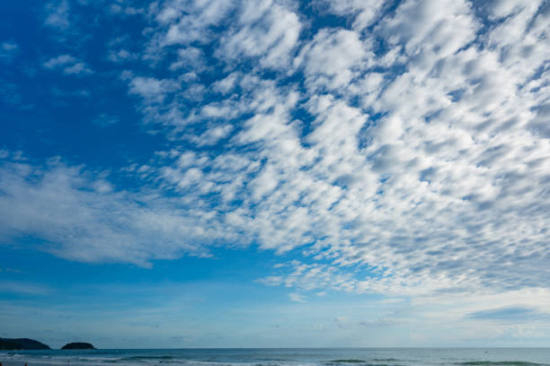 A wide ocean view beneath a deep blue sky dotted with soft, scattered cumulus clouds. The turquoise waters of the Atlantic Ocean stretch calmly toward the horizon, with gentle waves forming subtle white lines near the shore. The composition emphasizes open space, balance, and clarity, highlighting the contrast between the textured cloud formations and the smooth expanse of sea.
