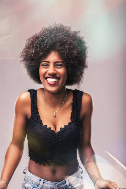 Portrait of a cheerful young woman with curly afro hair, beaming with a big smile while wearing a stylish black top and jeans, engaging directly with the camera
