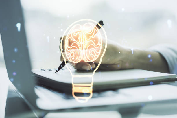 istock Creative light bulb with human brain hologram and hand writing in notepad on background with laptop, artificial Intelligence and neural networks concept. Multiexposure 2190159090