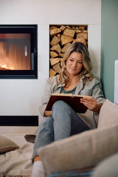 istock Smiling woman reading book while relaxing by fireplace at home. 2190168979