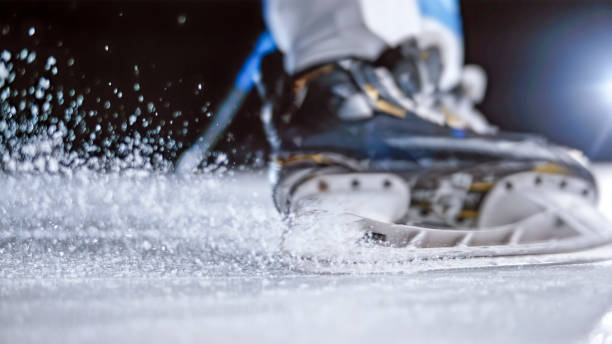 Low section of an ice hockey skates making a sharp turn across the rink, capturing the dynamic motion with ice spray and blade detail under dramatic lighting.