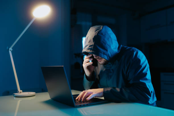 istock Hacker Typing on Laptop and Talking on Phone in Dark Room at Home 2190180746