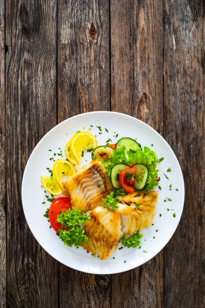 istock Fried codfish loin and fresh vegetables served on white plate on wooden table. 2190189247